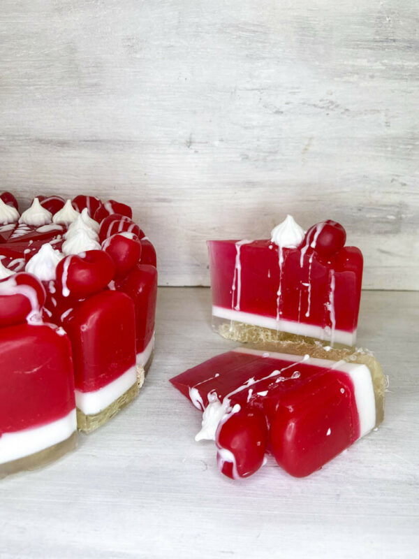 Χειροποίητη τούρτα σαπουνιού σε μορφή cheesecake με αποχρώσεις κερασιού και λευκού, διακοσμημένη με ρεαλιστικά στοιχεία και έντονο άρωμα κερασιού.